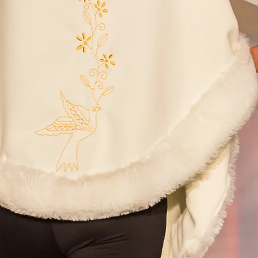 Detalhe da bainha de uma capa creme a cair, com um pássaro bordado a dourado